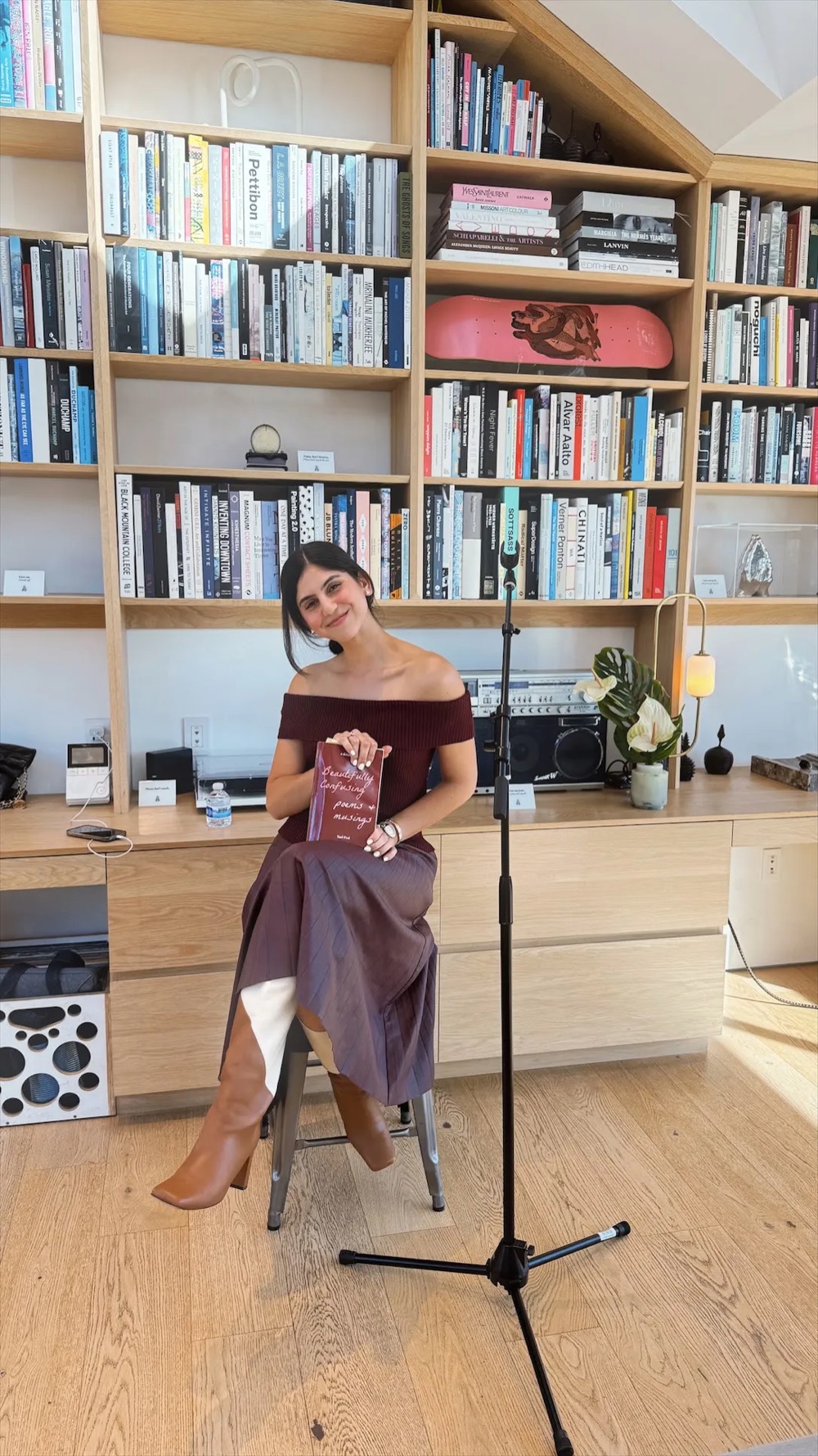 Yael Frai seated on a stool in front of tall bookshelves, smiling and holding a copy of Beautifully Confusing.